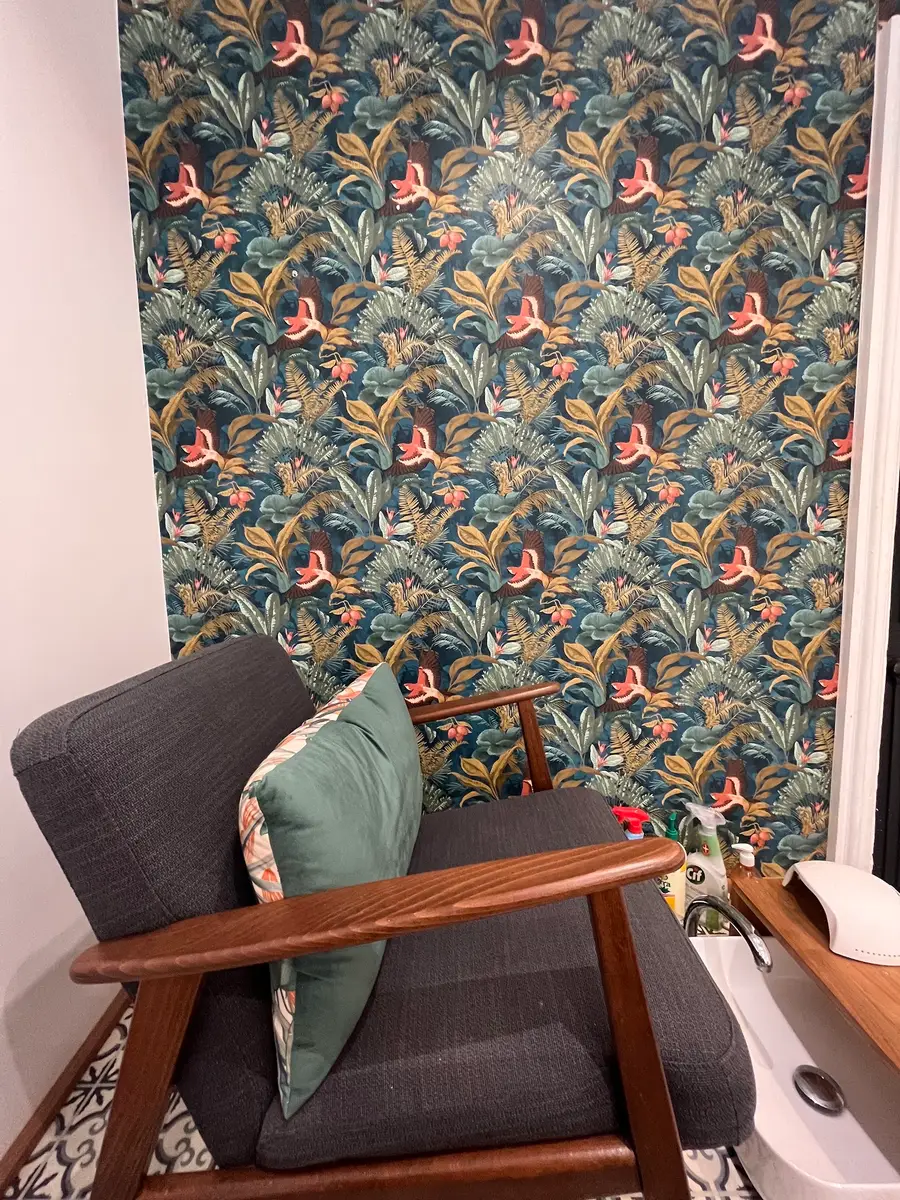 Fauteuil en bois et tissu gris devant un papier peint tropical, bac pour les pieds à droite