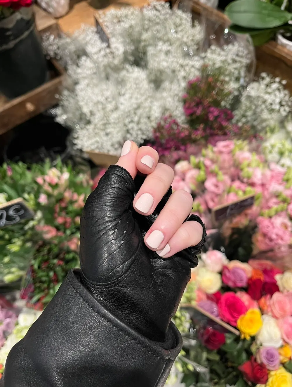 Main gantée de cuir aux ongles rose poudré devant des fleurs coupées