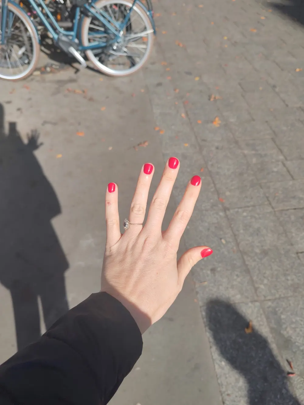 Ongles courts vernis rouge vif, main levée au-dessus d’un trottoir parisien