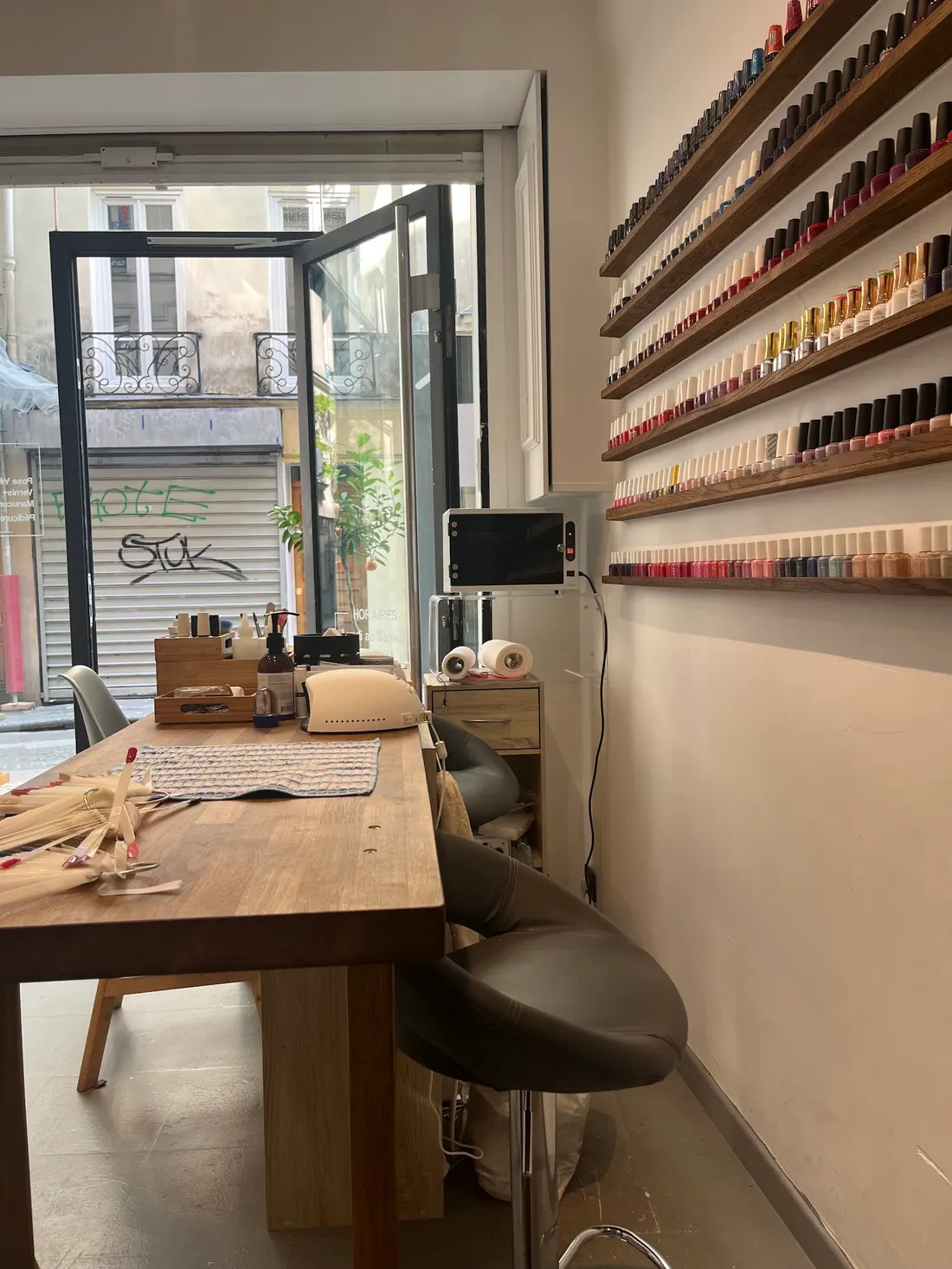 Intérieur du salon : table en bois face à la porte vitrée, étagères de vernis sur le mur blanc