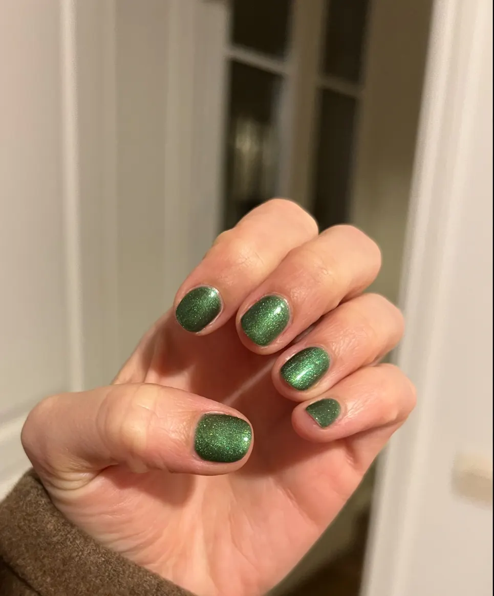 Ongles courts arrondis en vert pailleté, main repliée près d’une porte blanche
